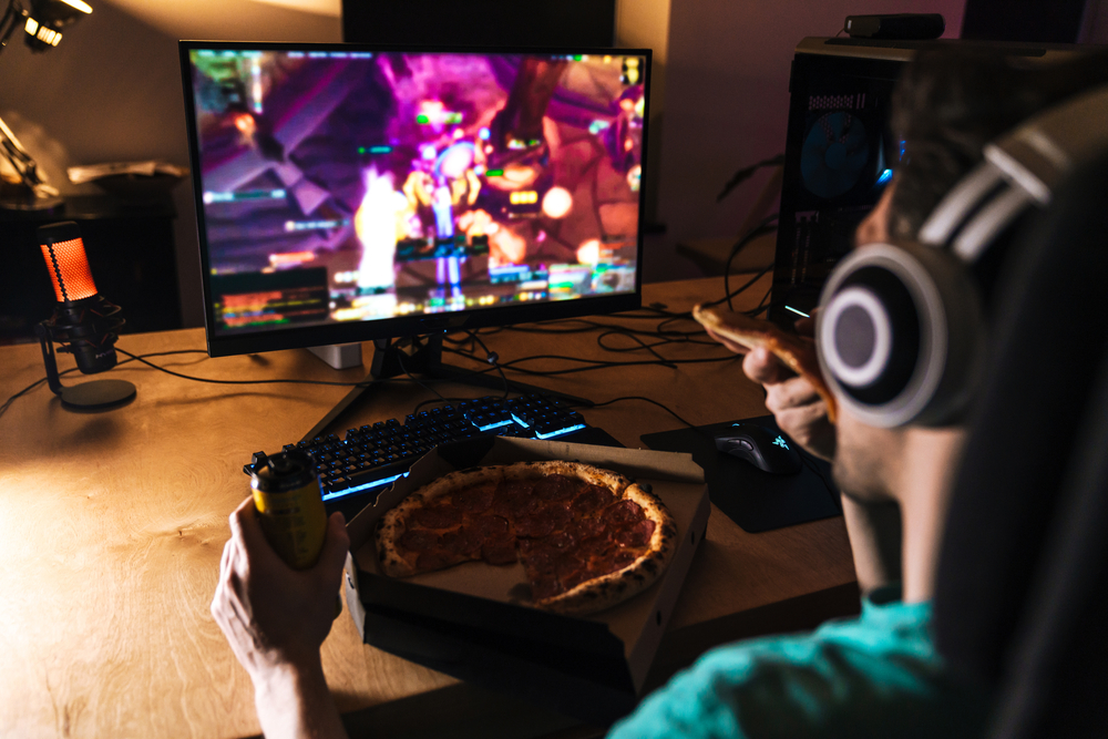 Image of focused young man eating pizza while playing video game